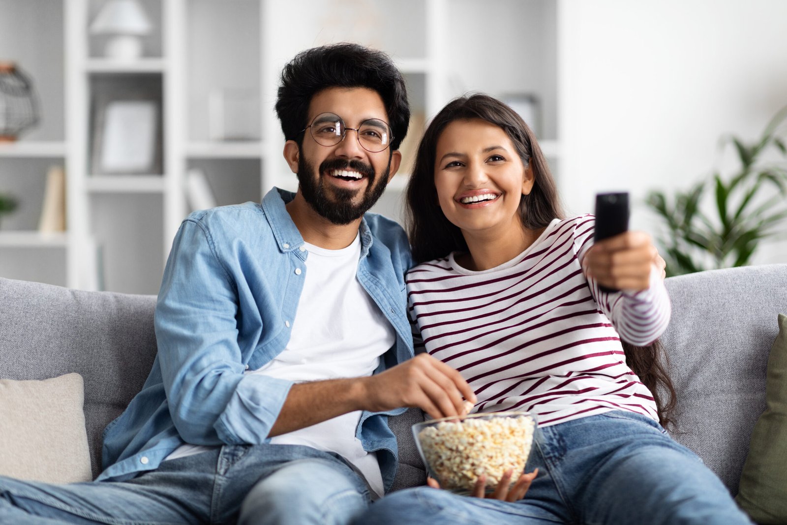 Smiling interracial spouses sitting on couch at home, watching tv Smiling attractive millennial interracial spouses middle eastern man and insian woman in casual sitting on couch at home, hugging, watching tv, eating popcorn, enjoying favorite series or show